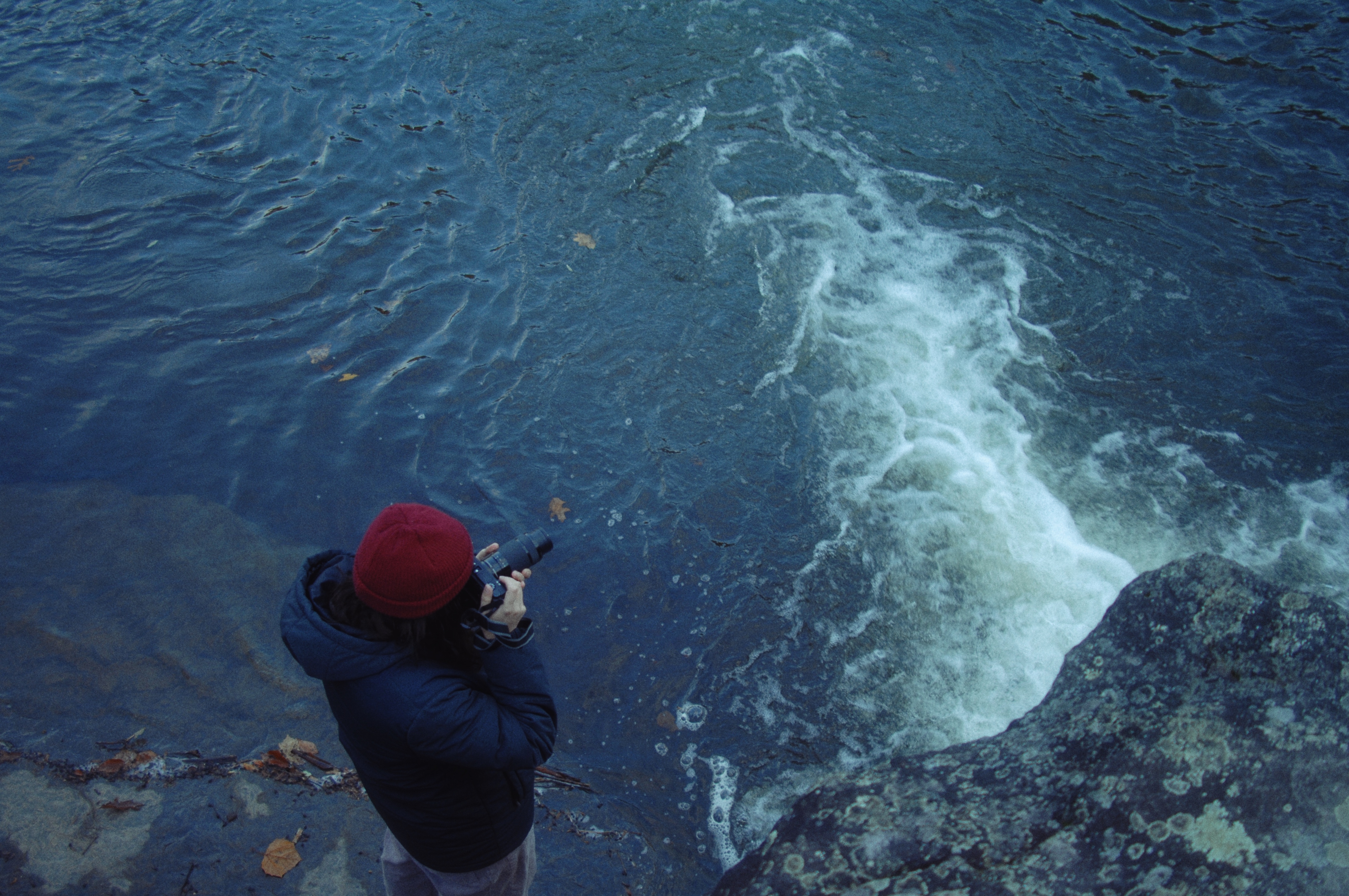 A person looking through the lens of a camera standing next to whitewater rapids.
