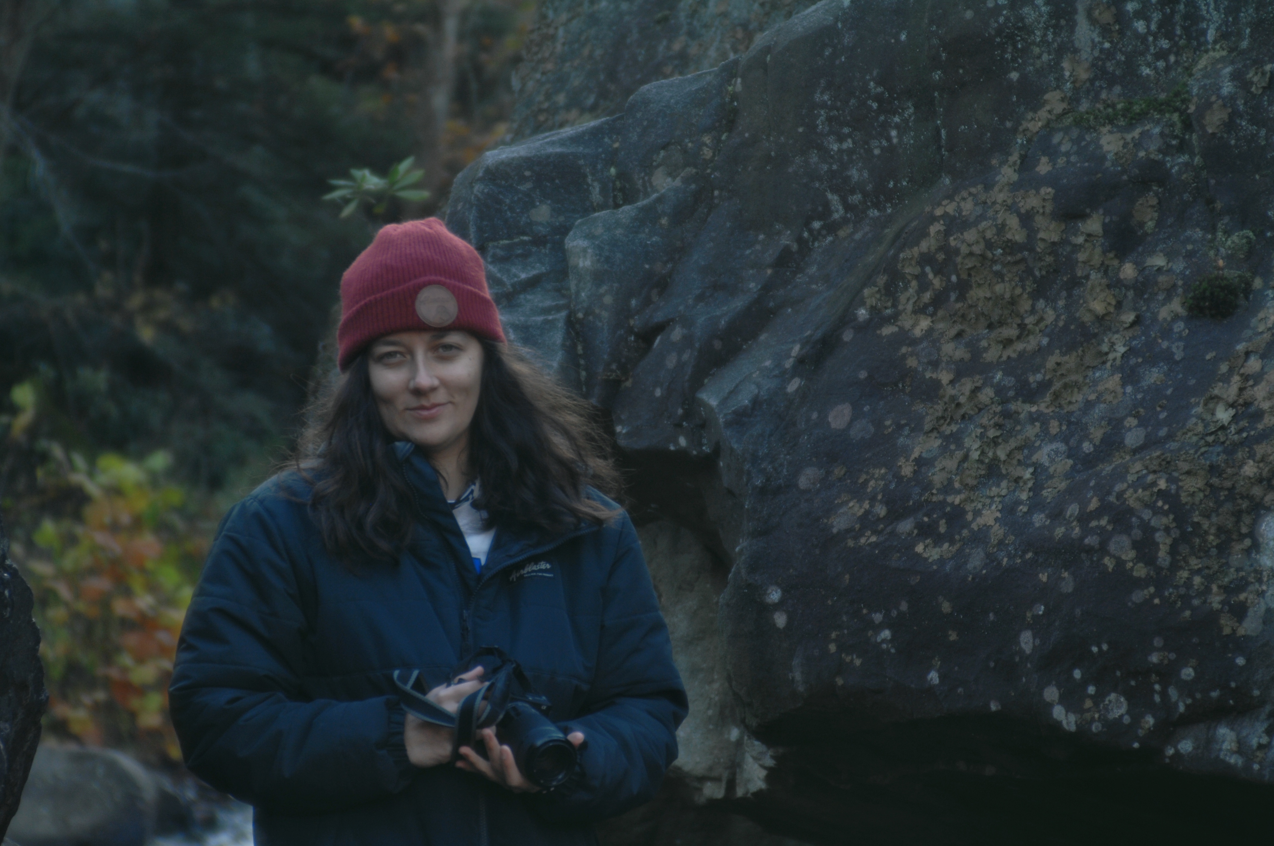 A portrait of a woman wearing a red hat and a black jacket holding a camera and standing in front of a large rock.